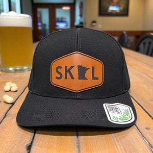 May include: Black baseball cap with a brown leather patch featuring the word "SKOL" and a silhouette of Minnesota. The cap has a curved brim and a white tag with a QR code.