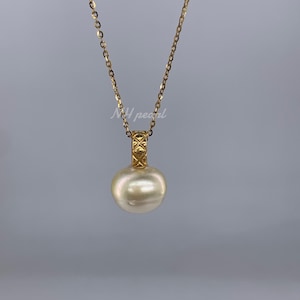 Puede incluir: Un collar dorado con un gran colgante de perla redonda de color crema. El colgante está suspendido de una fianza dorada decorativa. La cadena es delicada, sobre un fondo gris liso.