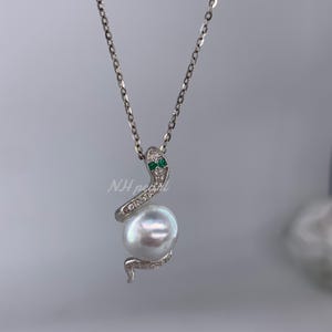 May include: A silver necklace with a snake pendant. The pendant has small, clear stones and two green stones, holding a large, white pearl. The chain is delicate, creating an elegant design. The necklace is suitable for special occasions.