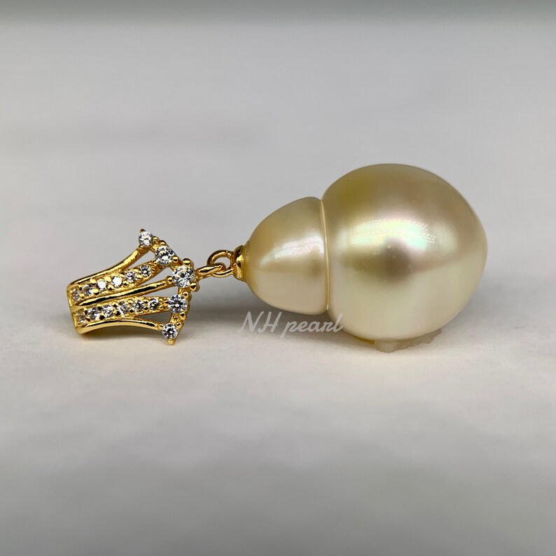 Puede incluir: Un colgante de perla barroca grande, de color blanco roto, con engaste de oro y piedras de circonita c&uacute;bica transparentes. La perla tiene un ligero brillo iridiscente.