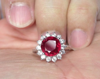 Puede incluir: Un anillo de plata con una gran piedra de rubí roja redonda rodeada por un halo de diamantes pequeños y transparentes.