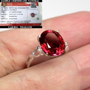 Może przedstawiać: Srebrny pierścionek z dużym, owalnym, purpurowo-czerwonym rubinem. Pierścionek osadzony jest w delikatnym, ozdobnym wzorze. Rubin ma wymiary około 10,52 mm x 8,47 mm x 5,92 mm i waży 2,71 grama.