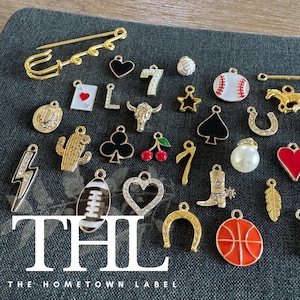 May include: A collection of gold-toned and enamel charms, including a horseshoe, heart, cactus, and a baseball. The charms are arranged on a dark gray fabric. The text "THL" and "THE HOMETOWN LABEL" are visible.