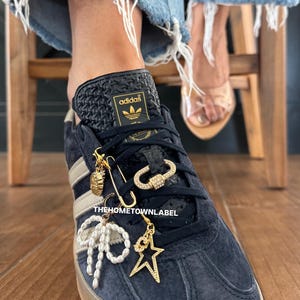 May include: A pair of black Adidas sneakers with gold accents and a variety of charms attached to the laces. The charms include a star, a pearl bow, a safety pin, and a gold chain with a diamond-encrusted clasp. The text "THEHOMETOWNLABEL" is printed on the laces.