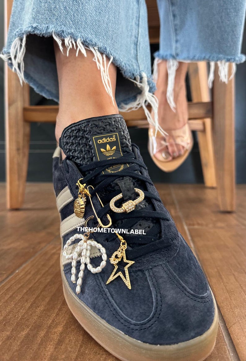 May include: A pair of black Adidas sneakers with gold accents and a variety of charms attached to the laces. The charms include a star, a pearl bow, a safety pin, and a gold chain with a diamond-encrusted clasp. The text "THEHOMETOWNLABEL" is printed on the laces.