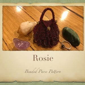 May include: A close-up of a beaded purse with a gold clasp. The purse is red and has a fringe. The purse is sitting on a wooden surface with a few stones.