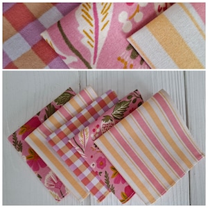 Può includere: Una collezione di tovaglioli di stoffa piegati in vari modelli e colori. I tovaglioli presentano disegni floreali, a righe e a quadri in tonalità di rosa, pesca e bianco. I tessuti sono disposti su una superficie di legno bianco.