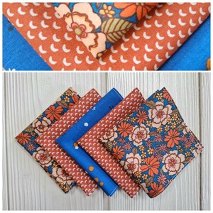 Può includere: Una collezione di sei fazzoletti di stoffa piegati. I fazzoletti presentano vari motivi, tra cui disegni floreali in arancione, blu e giallo, e un motivo a mezzaluna bianca ripetuta su uno sfondo color ruggine. Un fazzoletto è blu unito.