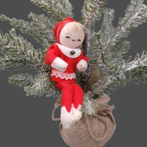 May include: A small doll with a round face, blonde hair, and a red felt outfit with white trim and lace detail, sitting atop a burlap-wrapped base. The doll is positioned in front of a flocked green pine branch with white snow-like accents.