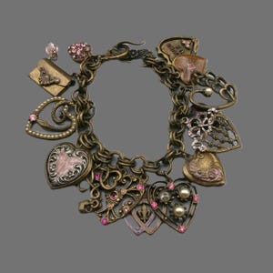 May include: A brass-toned charm bracelet featuring multiple heart-shaped pendants, intricate filigree metalwork, pink rhinestones, and small white pearls. The charms include a small envelope, various openwork hearts, and a heart with the text LOVES ME.