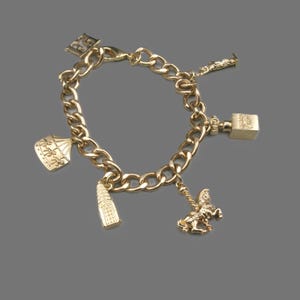 May include: A gold-tone charm bracelet featuring a thick curb chain and five distinct dangling charms, including a carousel, a tall building, a horse, a cube-shaped object, and a small figure, all finished in a polished metallic gold color.