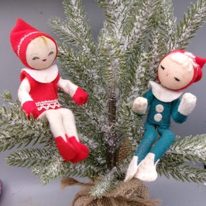 May include: Two vintage Christmas elf ornaments perched on a small, snow-covered evergreen tree. One elf wears a red outfit with white trim, the other a teal outfit. The tree is potted in a burlap-wrapped base, creating a festive holiday scene.