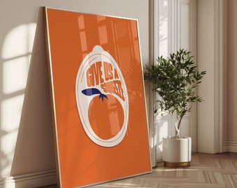 May include: An orange poster with a gold frame leans against a wall. The poster features a large white and blue graphic of a tomato with the text "GIVE US A SQUEEZE" in orange. The poster is next to a potted plant.