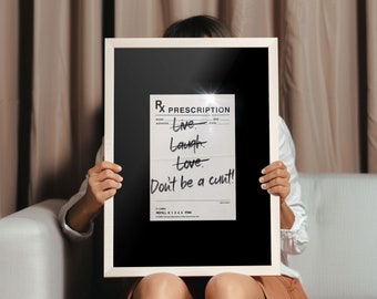 May include: A framed print featuring a prescription design with the words "Live, Laugh, Love, Don't be a cunt!" in handwritten style. The frame is white, and the background is black. The print is held by a person.