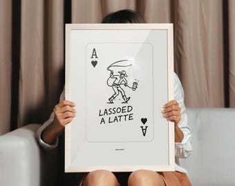 May include: A framed art print featuring a black and white illustration of a cowboy roping a coffee cup. The text "LASSOED A LATTE" is below the illustration. The artwork is designed to look like a playing card, with the Ace of Hearts displayed.