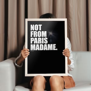 以下が含まれることがあります： 「NOT FROM PARIS MADAME.」というテキストが書かれた白黒のポスター。ポスターは白いフレームに額装され、人が持っている。