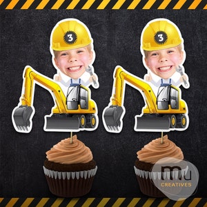 Op de afbeelding: Twee cupcake toppers met een lachend kind met een gele bouwhelm met het nummer 3. Elke topper heeft een geel graafmachine ontwerp. De cupcakes hebben bruine frosting.