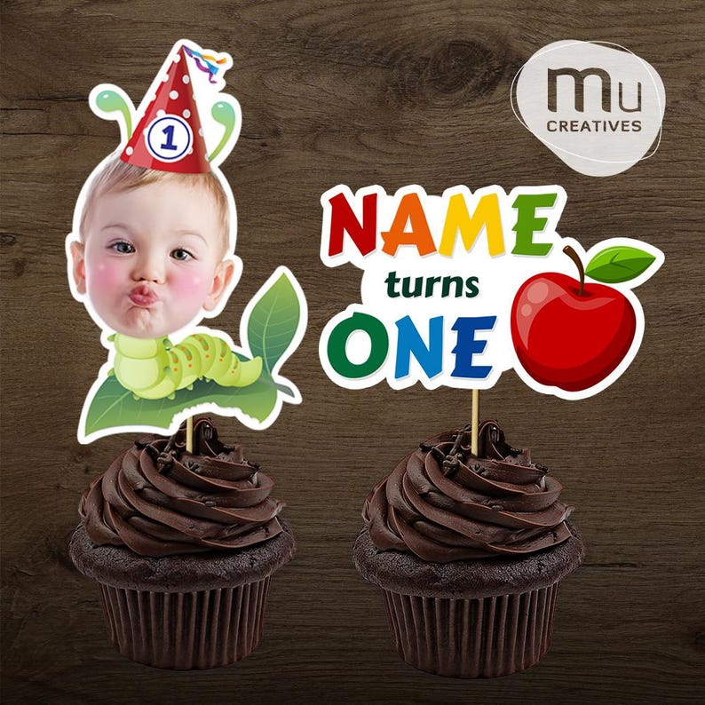 May include: Two chocolate cupcakes with decorative toppers. One topper features a baby's face with a party hat and a green caterpillar. The other says "NAME turns ONE" with a red apple.