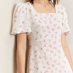 May include: A white dress featuring a square neckline and short, puffed sleeves. The dress is adorned with a pattern of small pink roses and green leaves. The text "suggestion of use by Opia Designs" is visible.