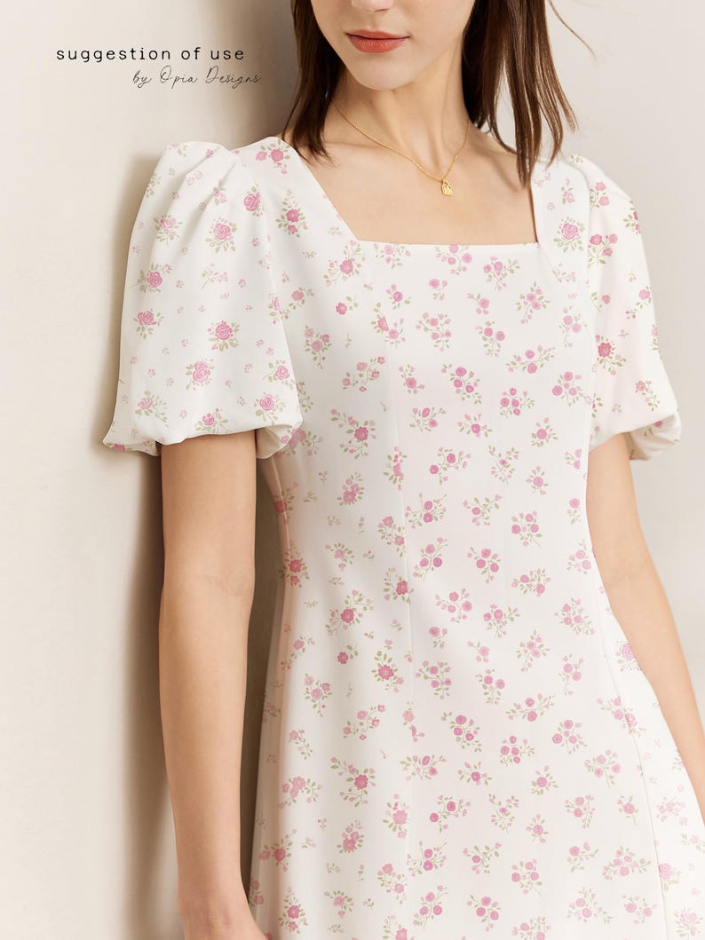 May include: A white dress featuring a square neckline and short, puffed sleeves. The dress is adorned with a pattern of small pink roses and green leaves. The text "suggestion of use by Opia Designs" is visible.