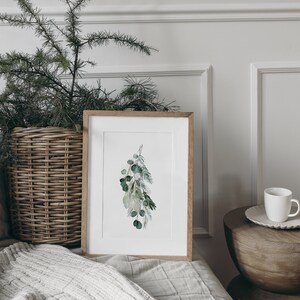 May include: A framed watercolor print of a sprig of green leaves hanging against a white wall. The print is in a light wood frame and is sitting on a white bed with a wicker basket and pillows.