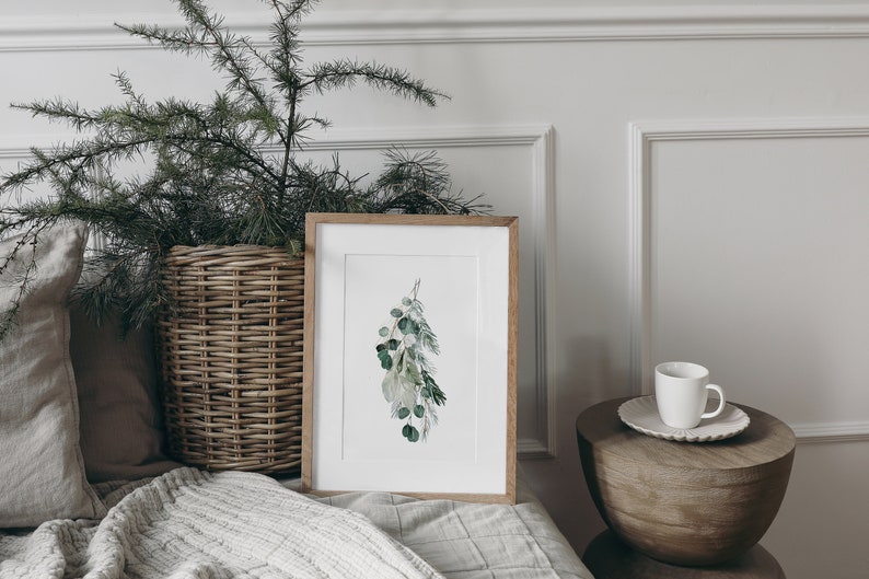 May include: A framed watercolor print of a sprig of green leaves hanging against a white wall. The print is in a light wood frame and is sitting on a white bed with a wicker basket and pillows.
