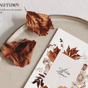 Op de afbeelding: Een aquarel-illustratie van herfstbladeren en bloemen op een witte kaart met de tekst "Herfstromantiek" op een kleinere witte kaart. De kaart ligt op een beige bord met twee bruine bladeren.