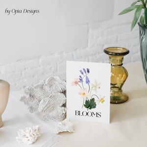May include: A white card featuring a watercolour illustration of a bouquet of flowers in shades of pink, blue, and white. The text "BLOOMS" is printed in black at the bottom of the card.
