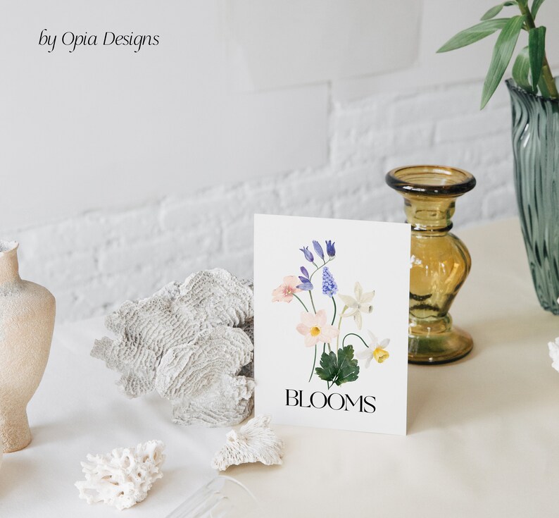 May include: A white card featuring a watercolour illustration of a bouquet of flowers in shades of pink, blue, and white. The text "BLOOMS" is printed in black at the bottom of the card.