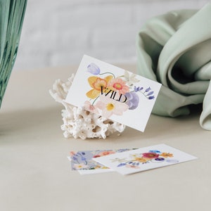 May include: A white card with a watercolour floral design in shades of pink, yellow, and blue. The word "WILD" is printed in black on the card. The card is propped up on a white coral-shaped object.