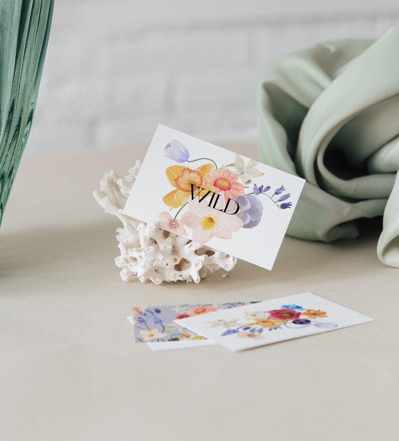 May include: A white card with a watercolour floral design in shades of pink, yellow, and blue. The word "WILD" is printed in black on the card. The card is propped up on a white coral-shaped object.