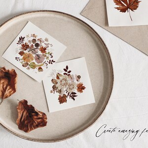 Op de afbeelding: Twee kaarten met gedroogde bloemenontwerpen op een beige bord. De kaarten bevatten een verscheidenheid aan gedroogde bloemen en bladeren in tinten bruin, roze en wit. De tekst "Create amazing fall designs" is in cursief geschreven op het witte oppervlak onder het bord.