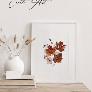 Op de afbeelding: Een wit frame met een aquarel schilderij van bruine bladeren en witte bloemen. Het frame staat op een houten tafel met een witte vaas en gedroogde grassen.