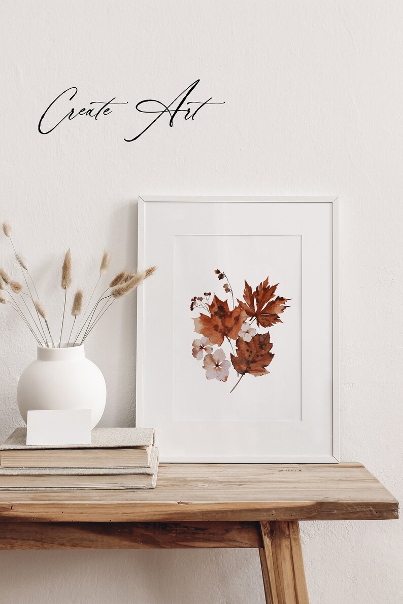 Op de afbeelding: Een wit frame met een aquarel schilderij van bruine bladeren en witte bloemen. Het frame staat op een houten tafel met een witte vaas en gedroogde grassen.