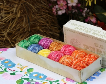 May include: A box of colorful embroidery floss in a white box with the text "D.O. PERLE" and "TRICOTON PARIS VILLAT" printed on the box. The box is sitting on a white tablecloth with a floral pattern.