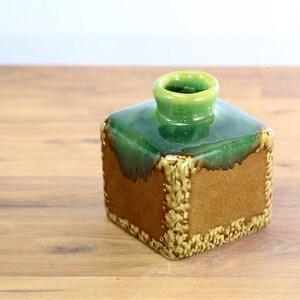 May include: A square ceramic vase with a green glaze and brown accents. The vase has a wide opening at the top and a textured surface.