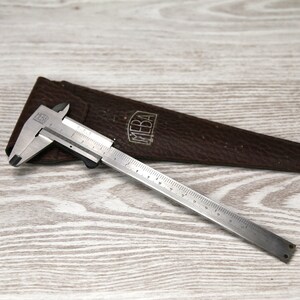 May include: A silver metal caliper with a brown leather case. The caliper has a ruler on the bottom and the case has the word "MEBA" embossed on it.