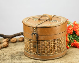 May include: A round, brown wicker and leather storage box with a handle on top. The box is sitting on a burlap surface with a branch and walnuts.