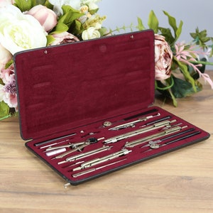 May include: A black leather case with a red velvet interior, open to reveal a set of drafting tools, including compasses, rulers, and pencils.