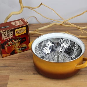 Op de afbeelding: Een ronde, mosterdgele geëmailleerde pan met twee handvatten, met daarin een inklapbare roestvrijstalen stoommand voor groenten. Naast de pan staat een bruine kartonnen doos met het label "STAINLESS STEEL VEGETABLE STEAMER".
