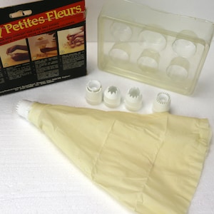 May include: A set of four white plastic piping tips and a beige piping bag. The tips are designed for making decorative flowers. The packaging is black with red and white text that reads "Classy Classy Petites Fleurs".