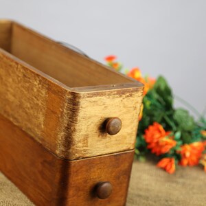 Peut inclure: Une bo&icirc;te en bois avec deux tiroirs, chacun avec un bouton rond. La bo&icirc;te est en bois clair et fonc&eacute; et a un aspect rustique. La bo&icirc;te est pos&eacute;e sur une surface en toile de jute.