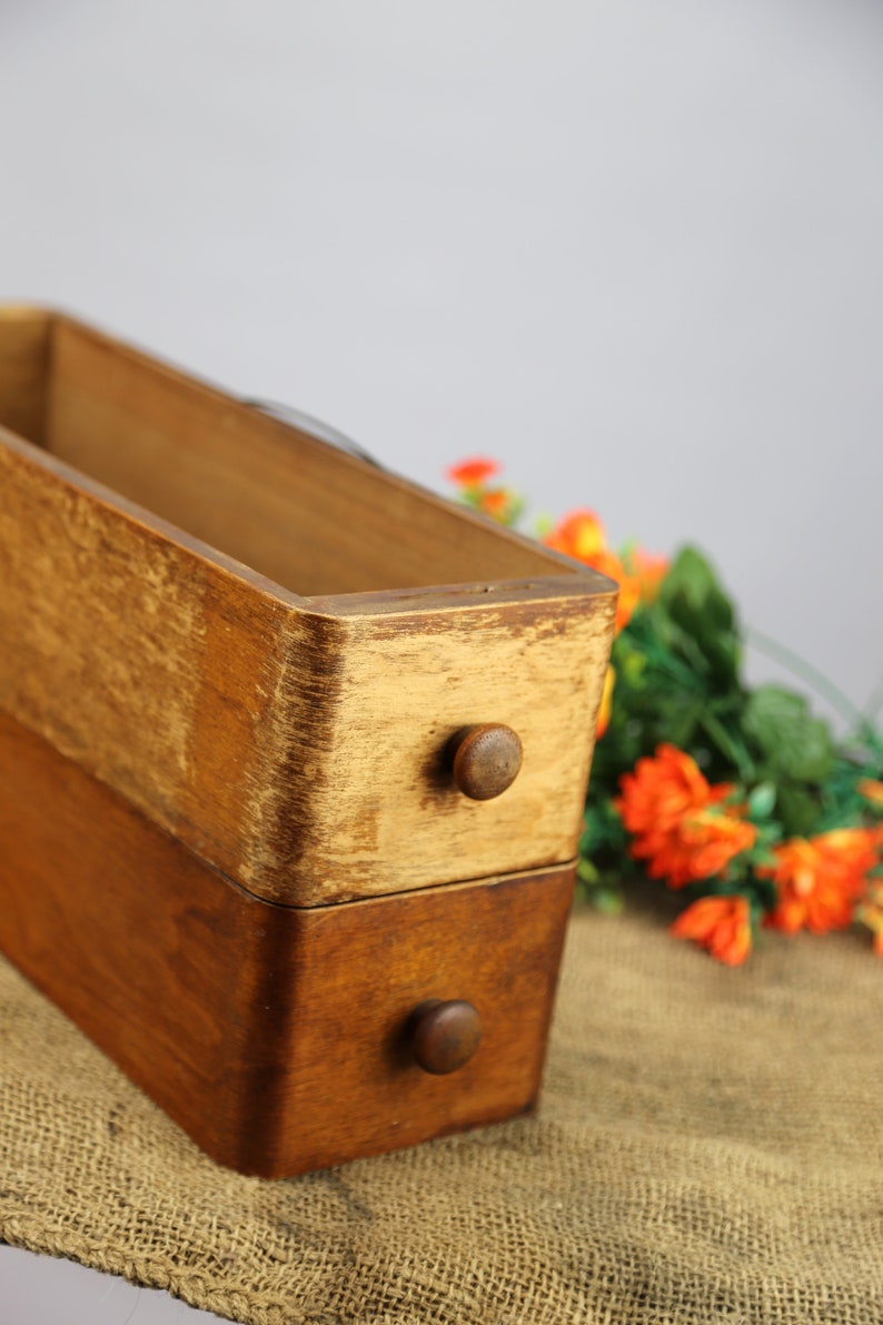 Peut inclure: Une bo&icirc;te en bois avec deux tiroirs, chacun avec un bouton rond. La bo&icirc;te est en bois clair et fonc&eacute; et a un aspect rustique. La bo&icirc;te est pos&eacute;e sur une surface en toile de jute.