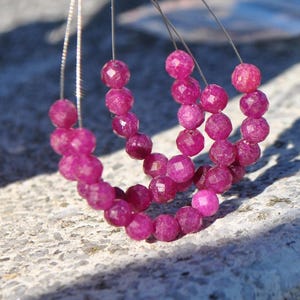 Peut inclure: Gros plan sur des perles de pierres précieuses rose foncé à facettes. Les perles sont enfilées sur un fil fin de couleur argentée et disposées en forme incurvée. La lumière du soleil crée des reflets et des ombres. L'arrière-plan est une surface texturée et claire.