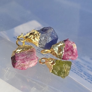 Puede incluir: Cuatro colgantes de piedras preciosas en bruto en tonos azul, rosa y verde, con tapas y bucles de metal dorado. Las piedras preciosas tienen una textura rugosa y natural. Los colgantes se muestran sobre un fondo azul claro.