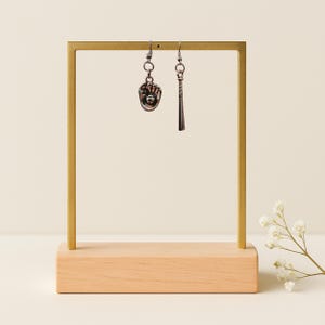 May include: A pair of silver-toned earrings hanging from a gold-colored metal stand with a light wood base. One earring is shaped like a baseball glove with a ball, and the other earring is shaped like a baseball bat.