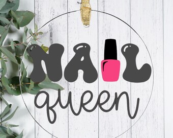 May include: A clear, round ornament with the words "NAIL QUEEN" in black and a pink nail polish bottle design. The ornament hangs from a gold ribbon against a white wood background.