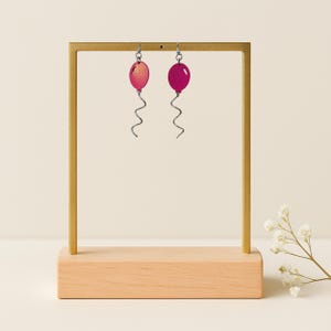May include: A pair of magenta balloon-shaped earrings with silver-tone wire details. The earrings are displayed on a gold-colored metal stand with a wooden base. The background is a neutral cream color.