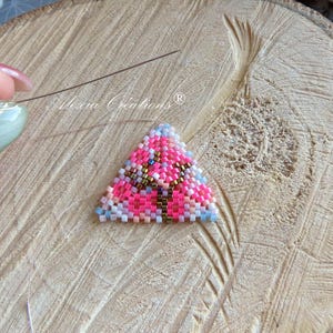 Può includere: Primo piano di un triangolo di perline in lavorazione, con perline rosa, blu e marroni. Il triangolo è su una superficie di legno. Vengono utilizzati ago e filo. Il testo "Alexia Creations" è visibile.