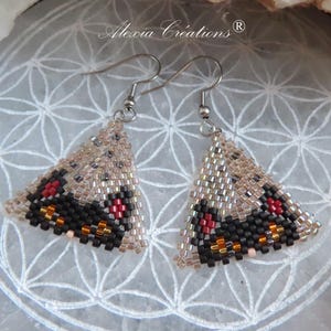 May include: Handmade beaded earrings in a triangular shape. The earrings feature a beige background with black, red, and orange accents. The earrings are suspended from silver-colored hooks. The brand name "Alexia Creations" is visible.